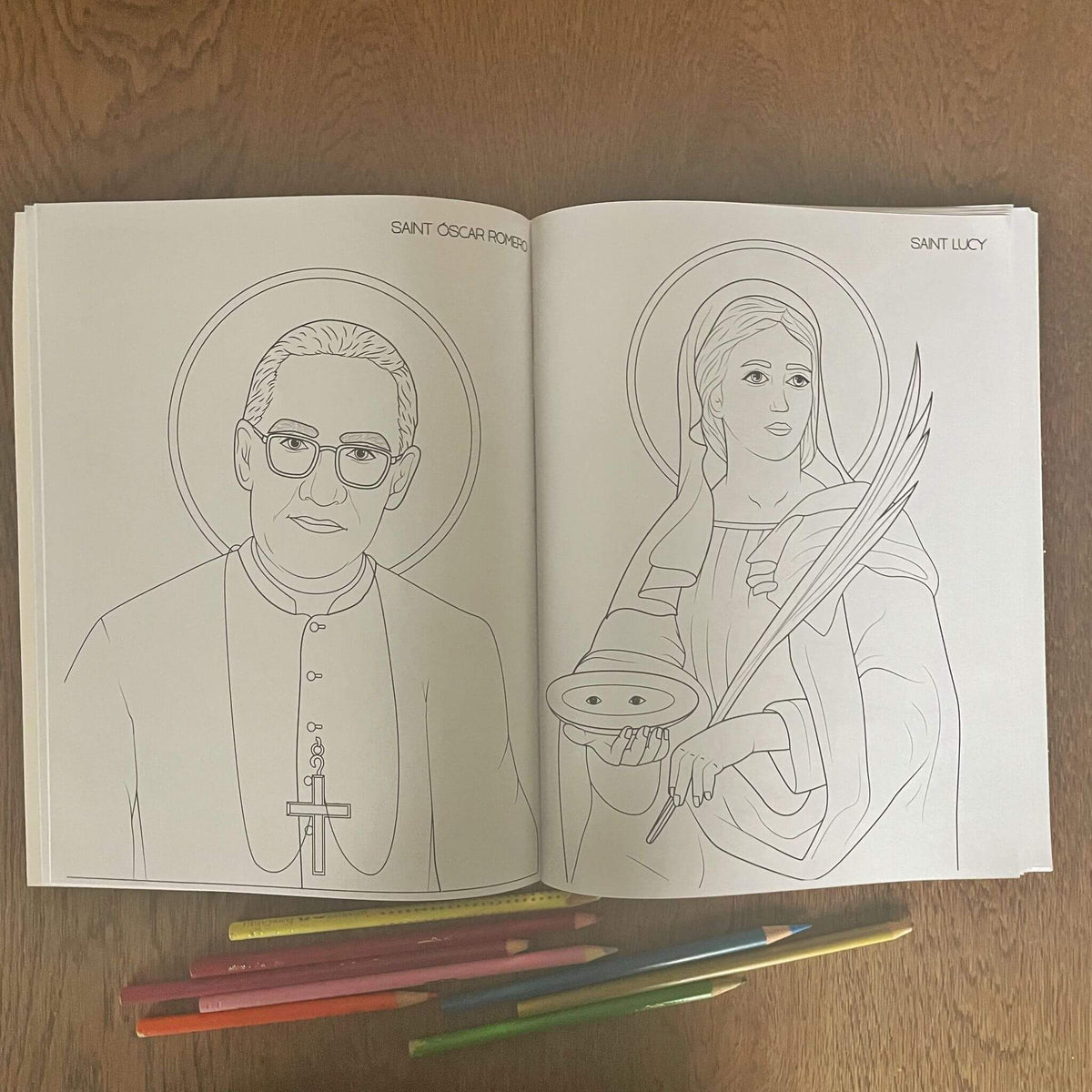 Coloring book open to pages of Saint Oscar Romero and Saint Lucy with colored pencils on table.