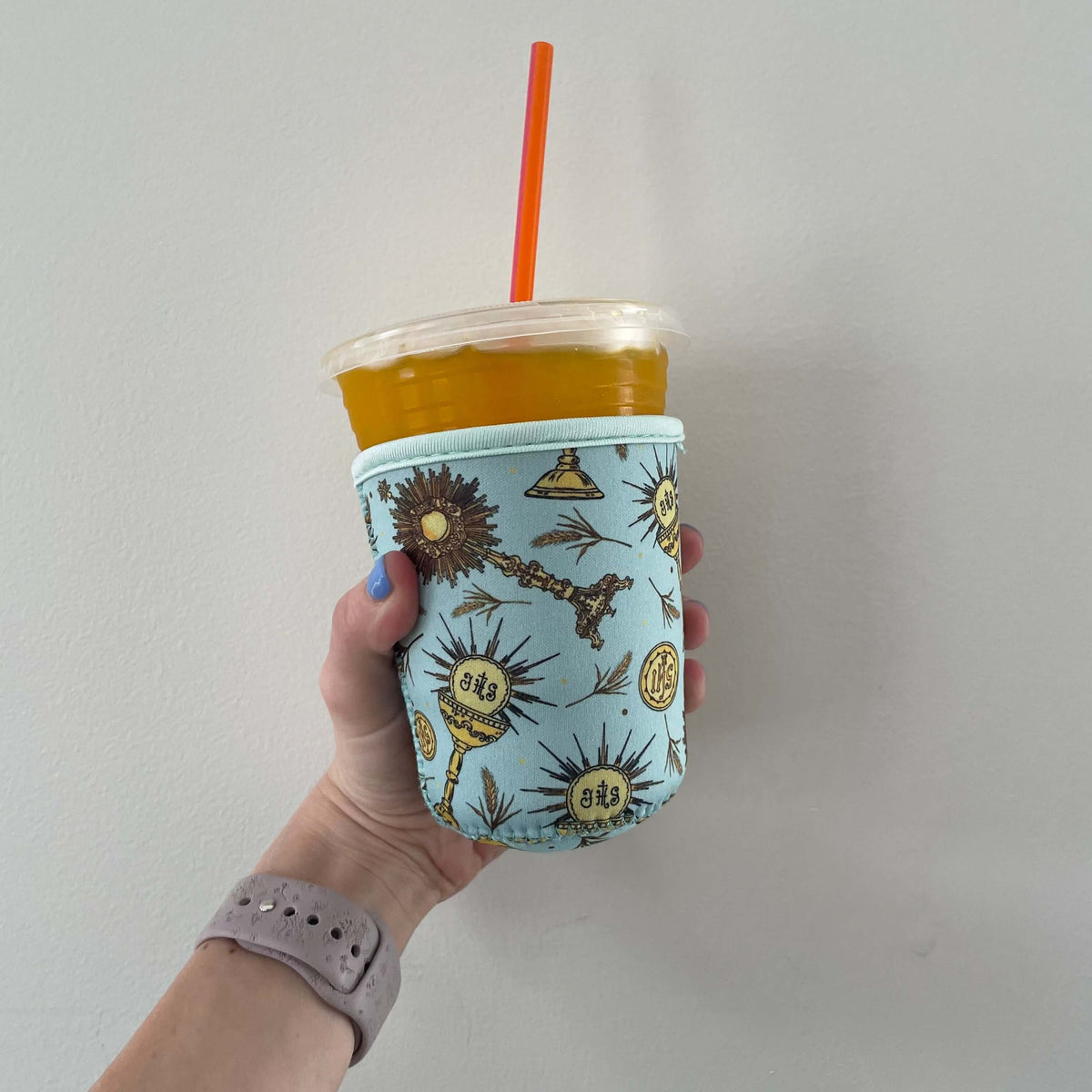 Holding a Catholic Cold Cup Sleeve with Eucharistic design, containing an iced drink, showcasing waterproof neoprene insulation.