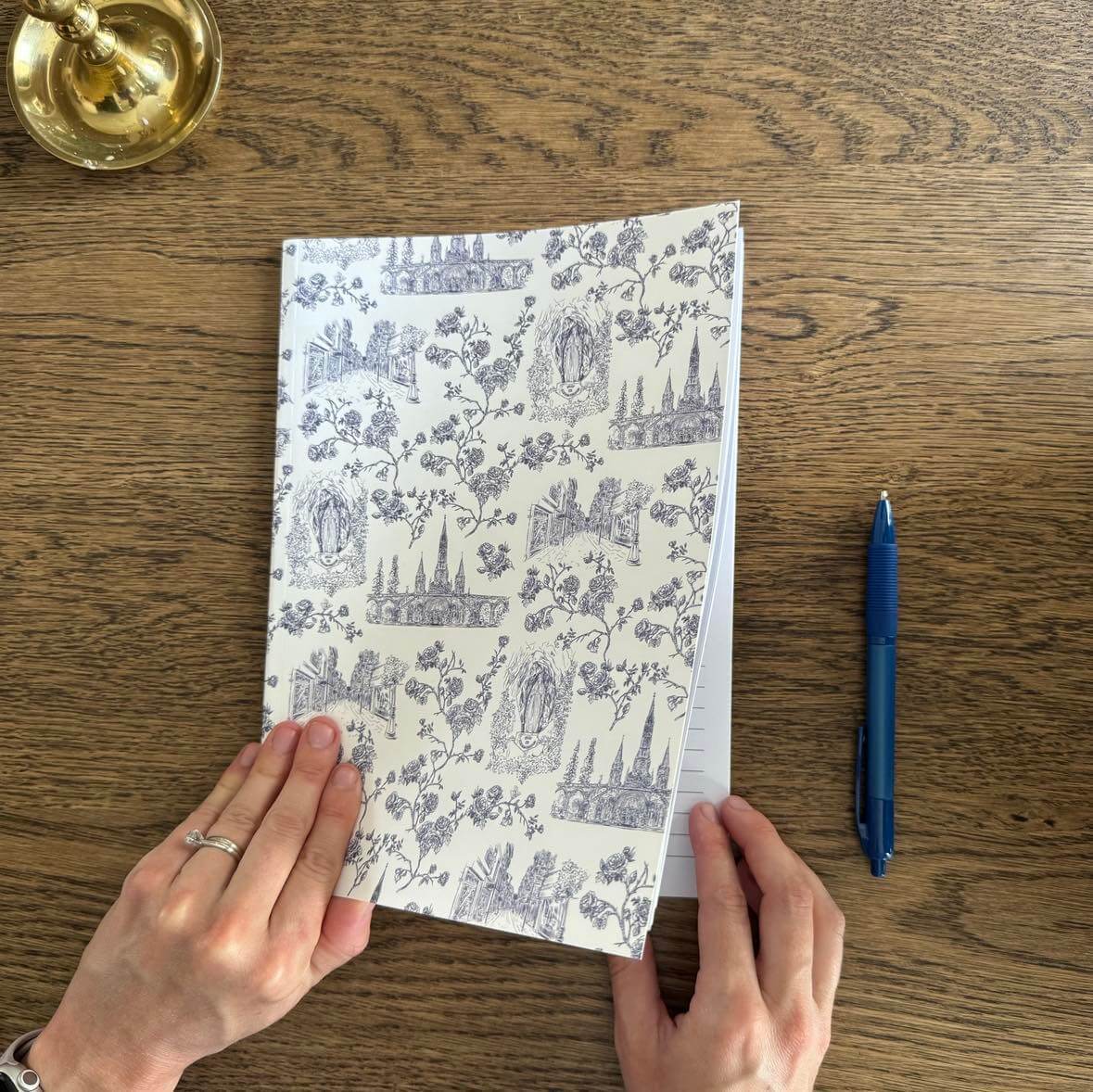 Alt Tag: Catholic Composition Notebook from Lourdes Collection on wooden table with pen, featuring intricate faith-themed design.