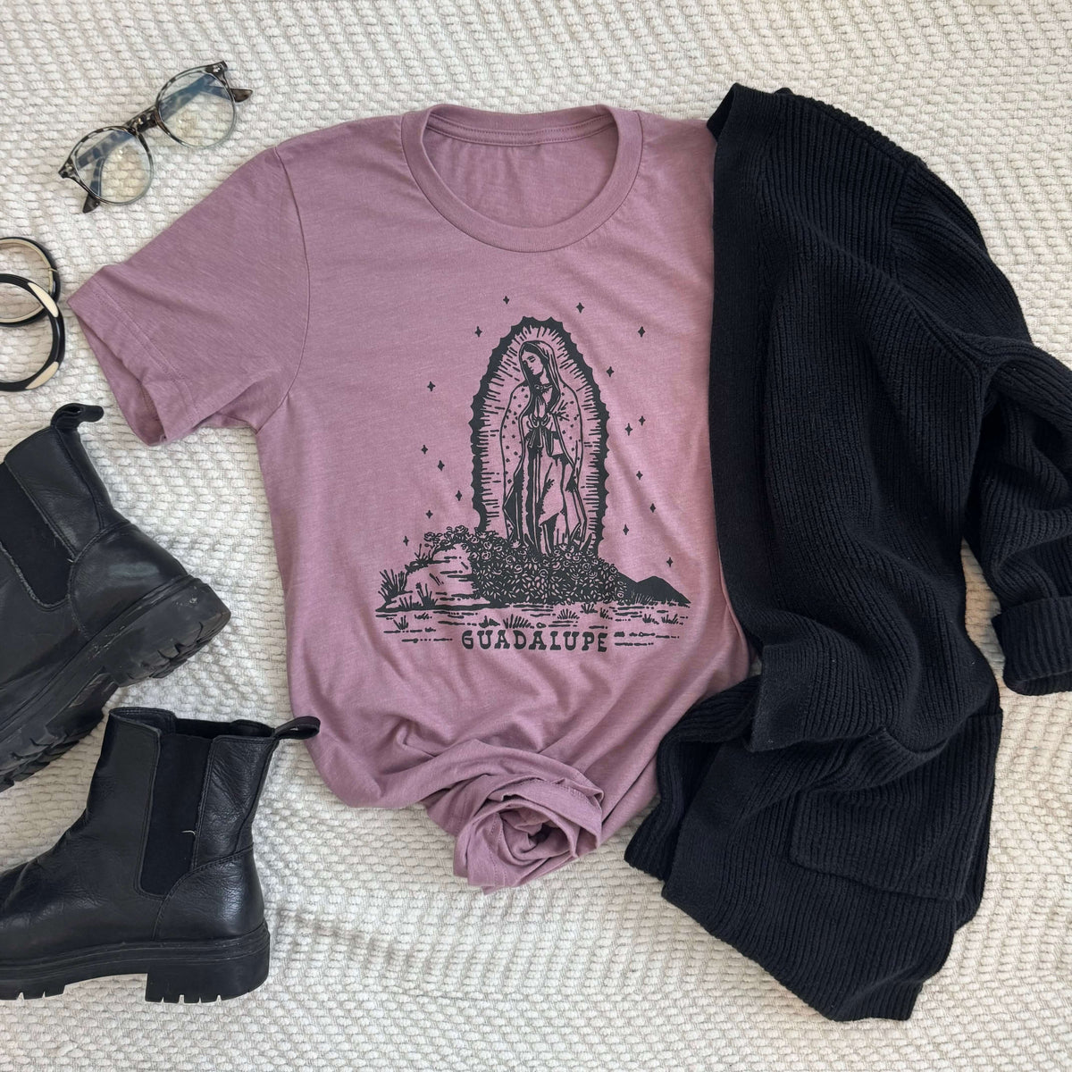 Our Lady of Guadalupe t-shirt displayed with boots, glasses, and a sweater on a textured surface; spiritual and stylish apparel.