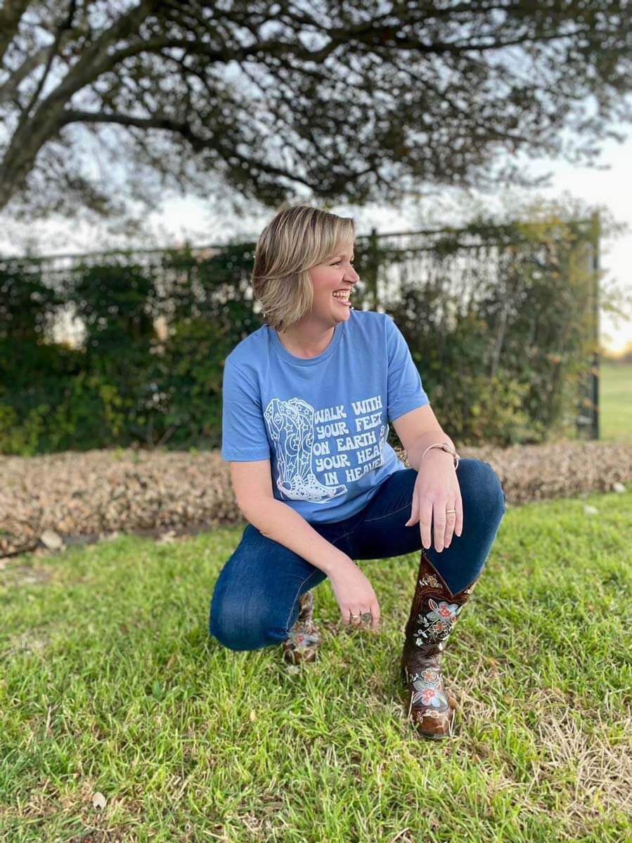 Woman wearing 'Walk With Your Feet On Earth' cowboy boots tee kneeling outdoors on grass.