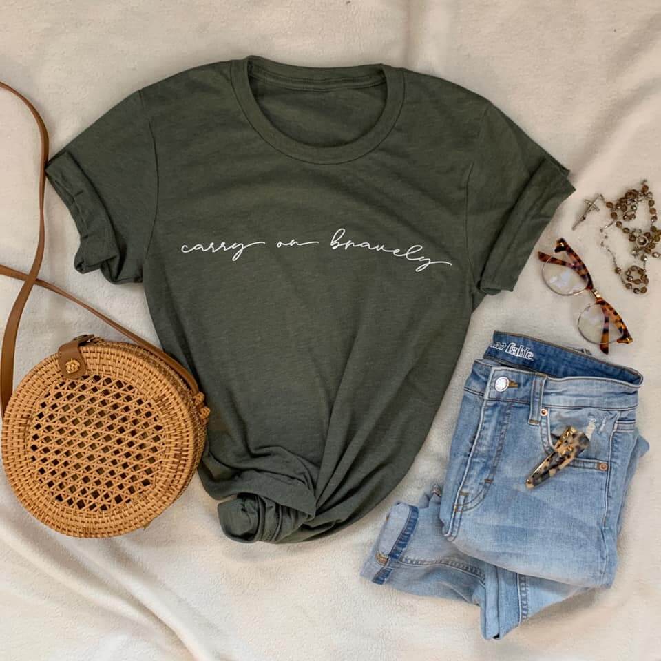 Moss green crew neck tee with 'carry on bravely' script, styled with a woven bag, denim shorts, sunglasses, and jewelry.