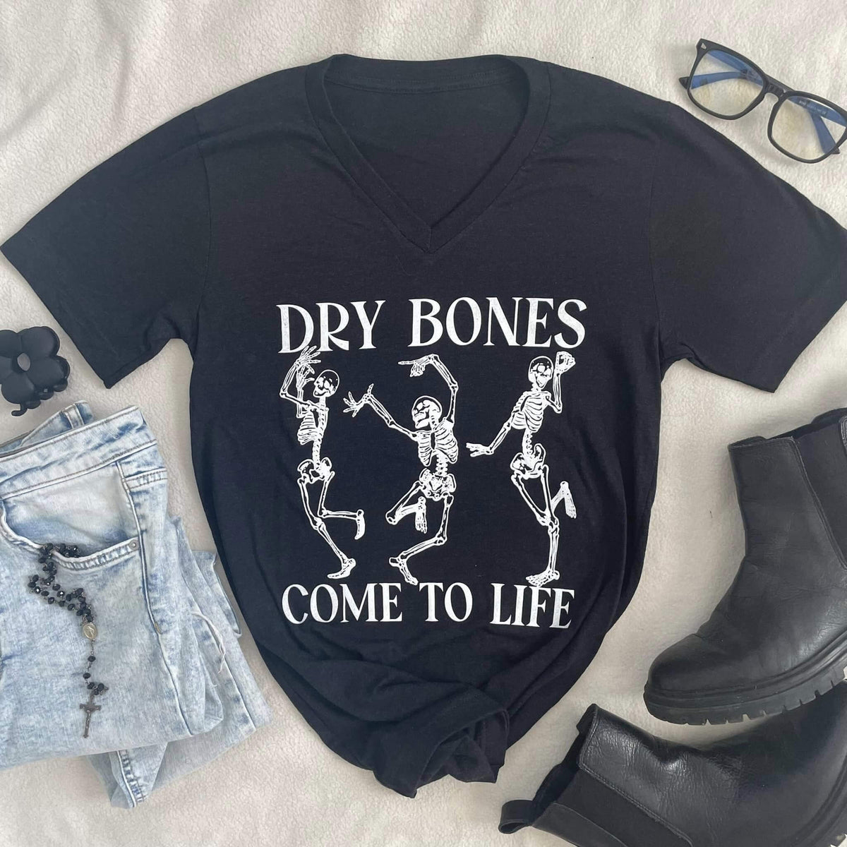 Black v-neck tee with 'Dry Bones Come to Life' text and skeleton print, paired with jeans, boots, glasses, and a bracelet.