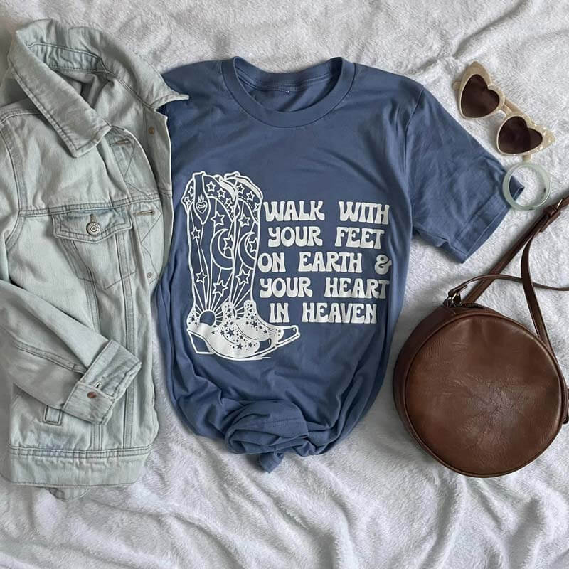 Graphic tee with cowboy boots design and quote 'Walk with your feet on Earth & your heart in Heaven', styled with denim jacket and sunglasses.