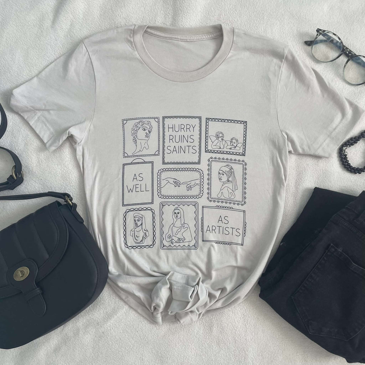 Heather dust crew neck tee with 'Hurry Ruins Saints' graphic, inspired by Thomas Merton, laid on a bed with handbag and accessories.
