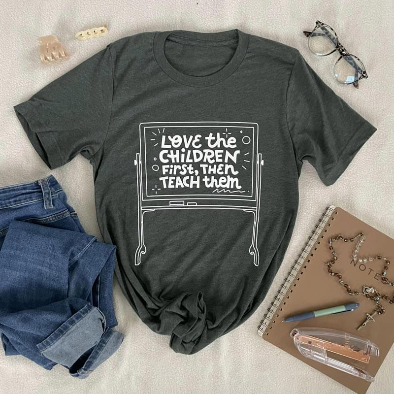 Dark green tee with "Love the children first, then teach them" text, inspired by Saint Mother Theodore Guerin, laid beside jeans and accessories.