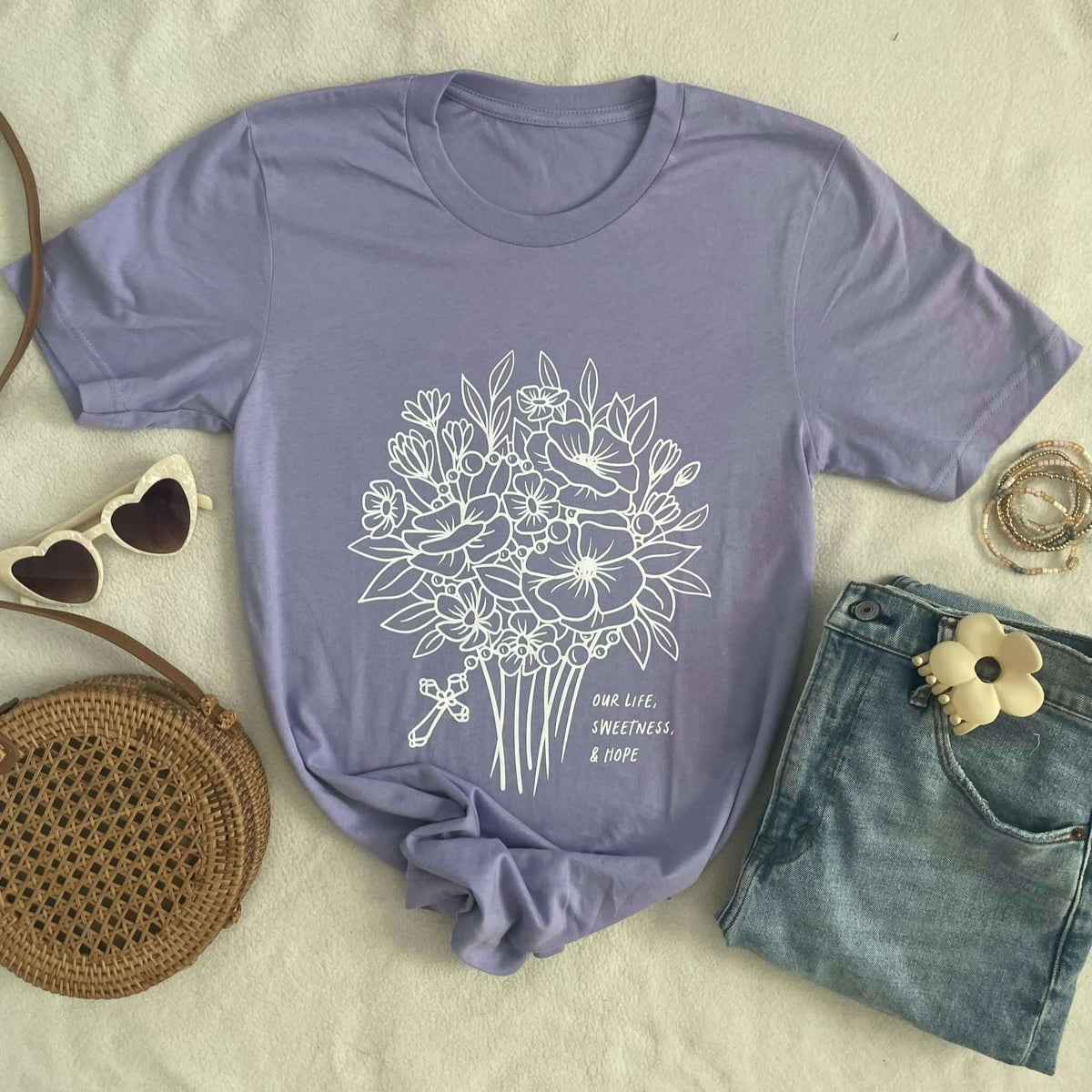 Lavender tee with floral design and text 'Our Life, Sweetness & Hope' styled with sunglasses and jeans on a soft surface.
