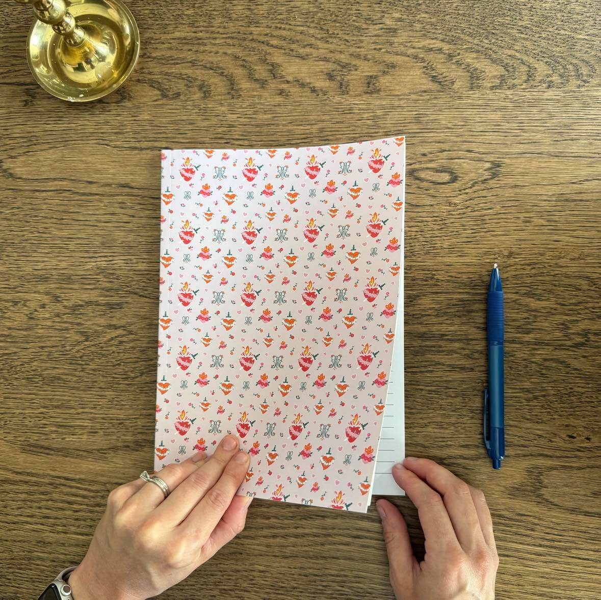 Auspice Maria B5 composition notebook on a wooden table with hands holding it, featuring religious-themed patterns and a blue pen beside it.
