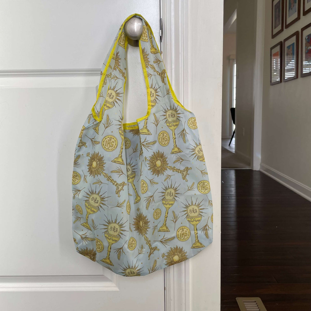 Catholic reusable grocery bag with Eucharistic design hanging on a door handle.