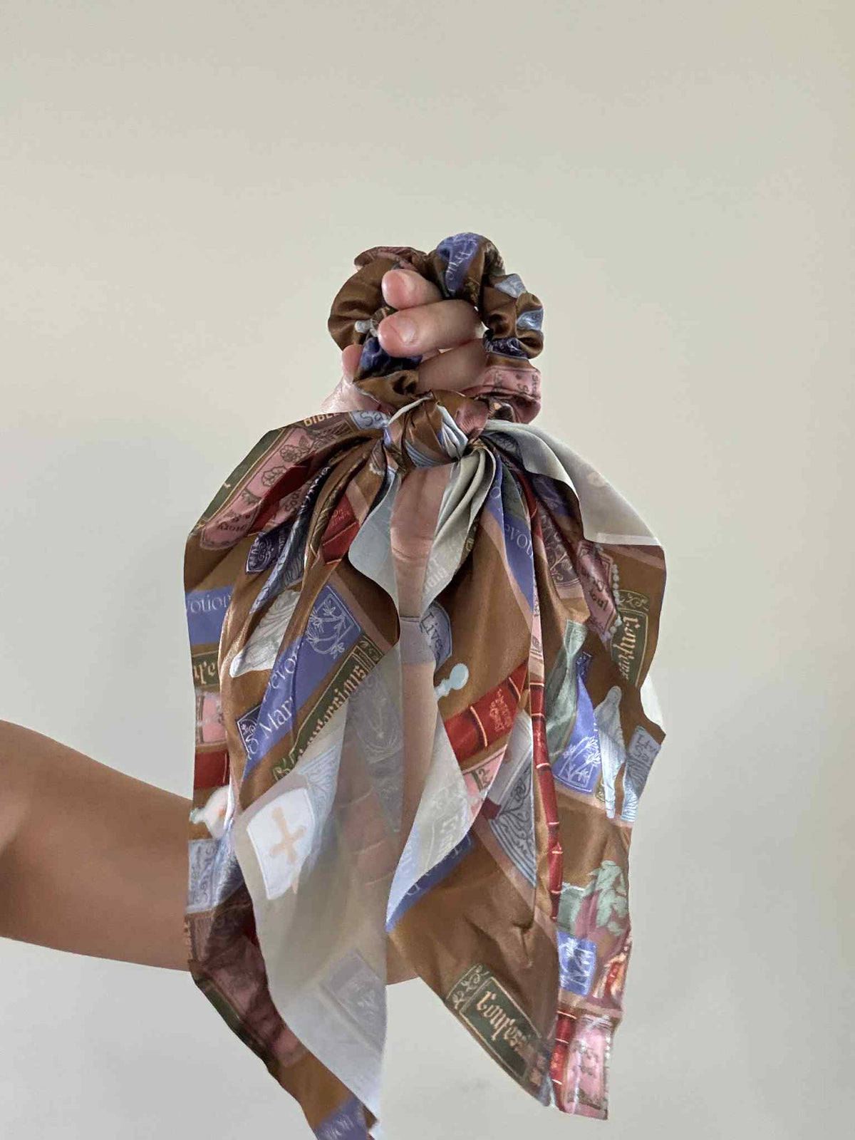 Hand holding a Catholic satin scrunchie with religious patterns, part of the Catholic Library Collection, combining faith and style.
