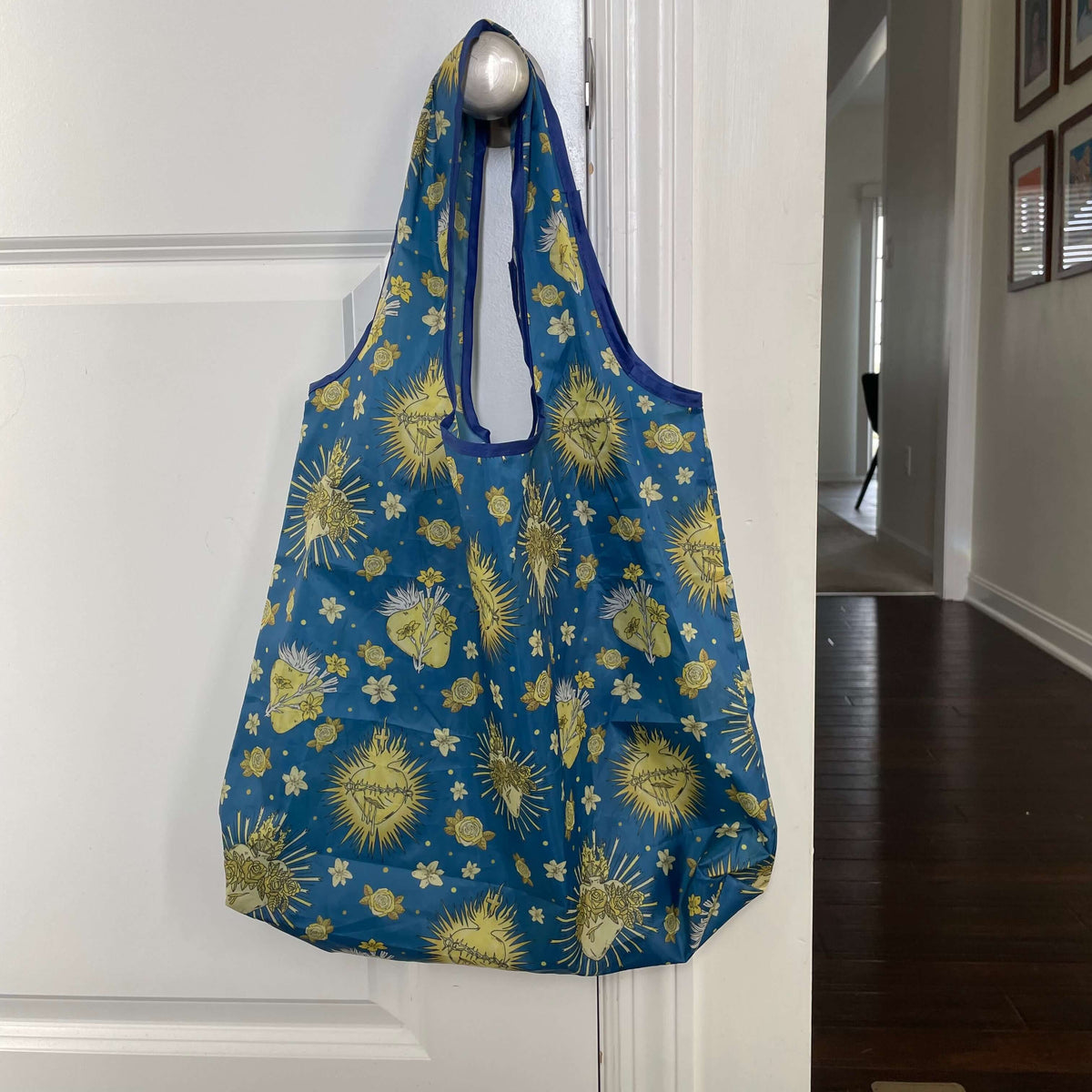 Holy Hearts Collection Catholic reusable grocery bag with vibrant religious pattern hanging on a door handle.