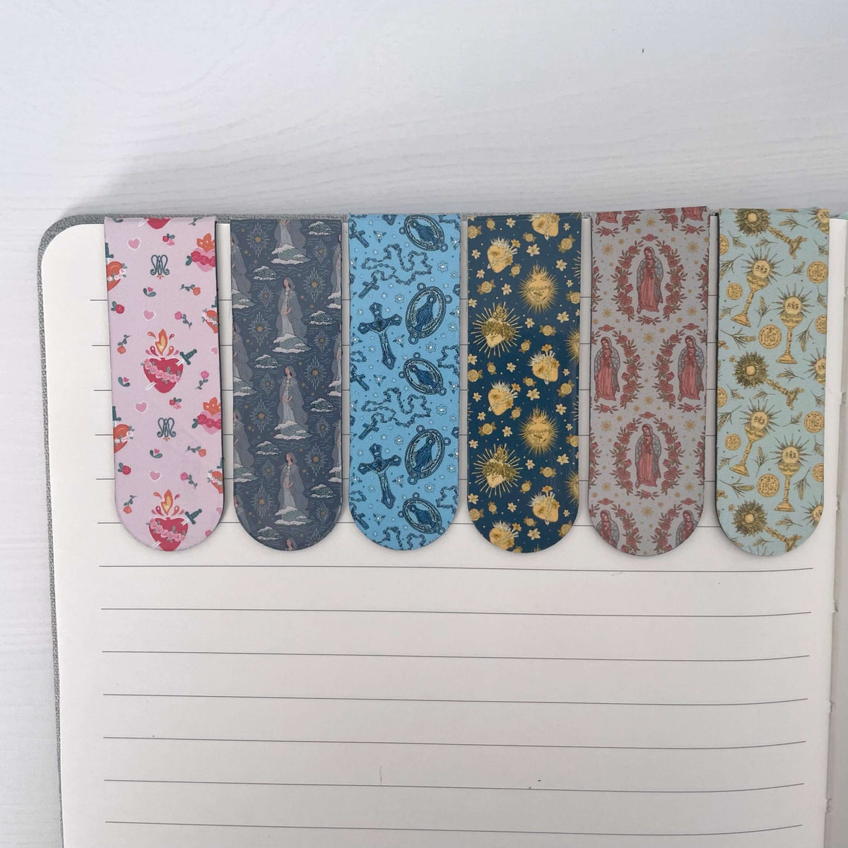 Set of 6 Catholic magnetic bookmarks with colorful sacred patterns on an open notebook page.