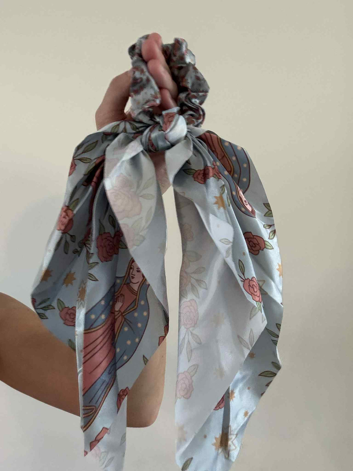 Hand holding a Catholic Satin Hanging Scrunchie from the Guadalupe Collection, featuring soft floral and religious patterns.