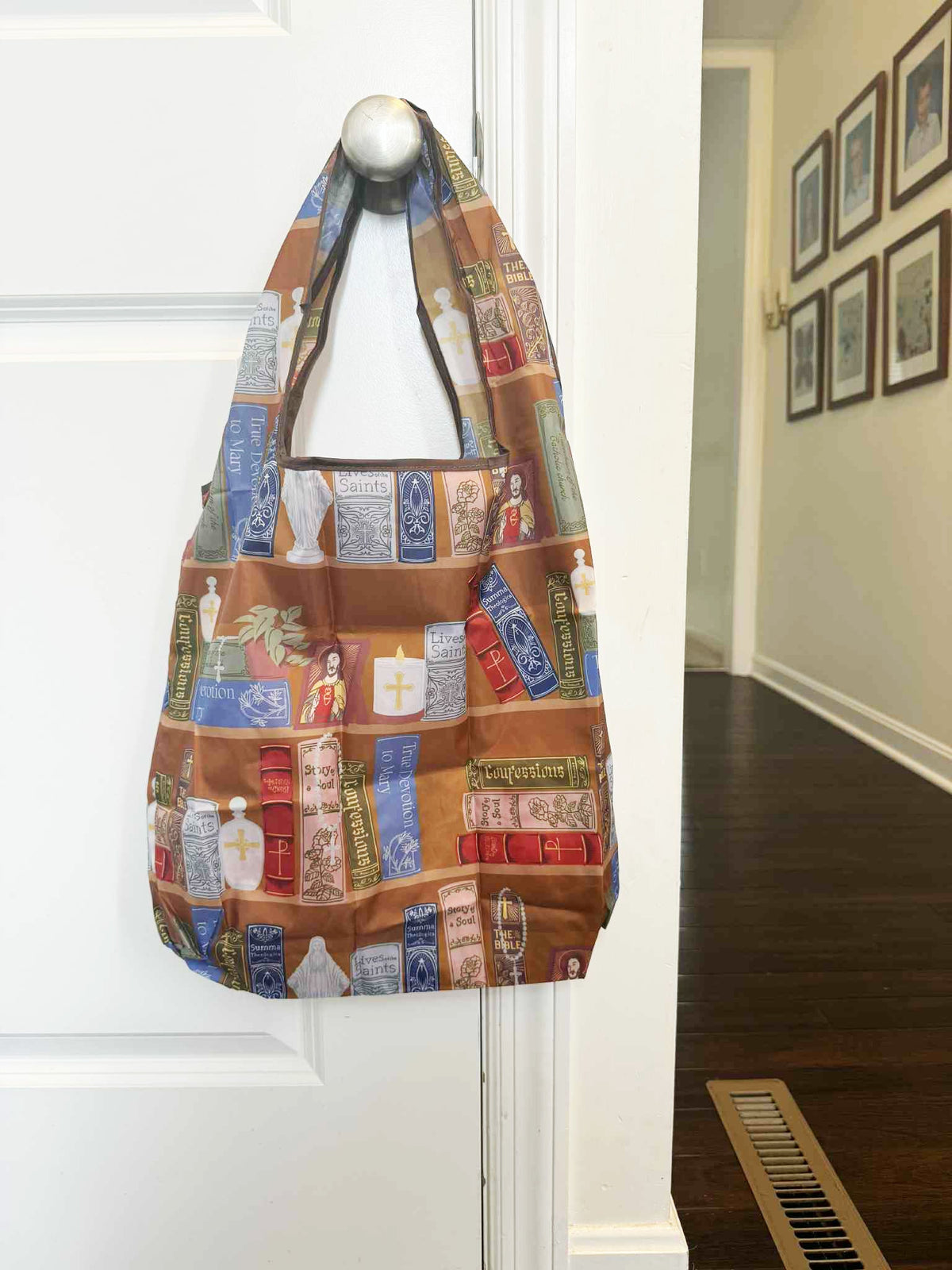 Catholic reusable grocery bag featuring religious patterns hung on a white door, promoting eco-friendly lifestyle.