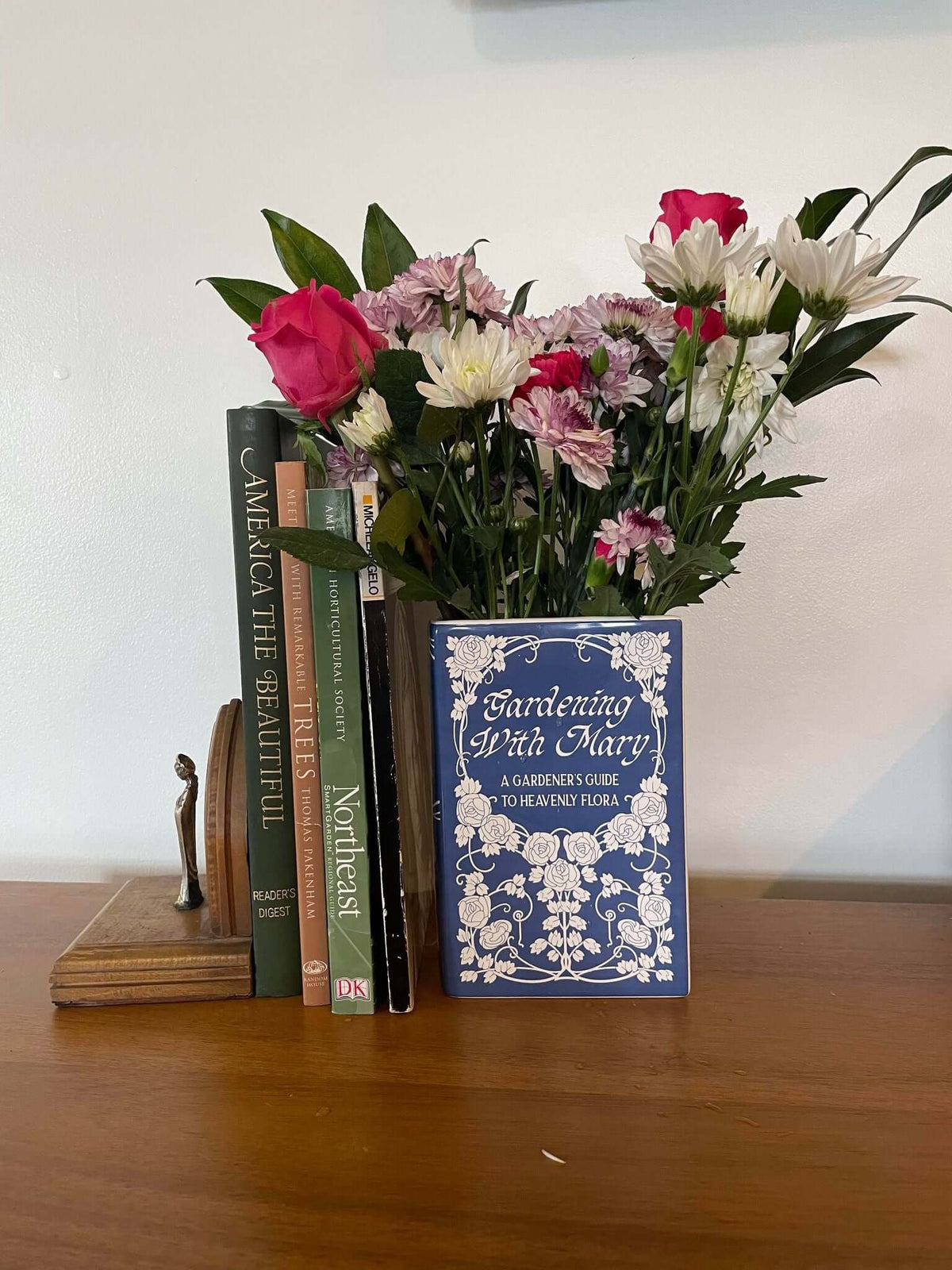 Ceramic book vase titled 'Gardening With Mary: A Gardener’s Guide to Heavenly Flora' with fresh flowers on a wooden shelf.