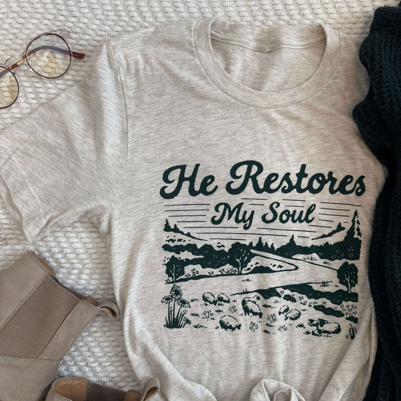 Alt Tag: He Restores My Soul t-shirt inspired by Psalm 23, featuring a peaceful scenic design, displayed on a cozy textile background.