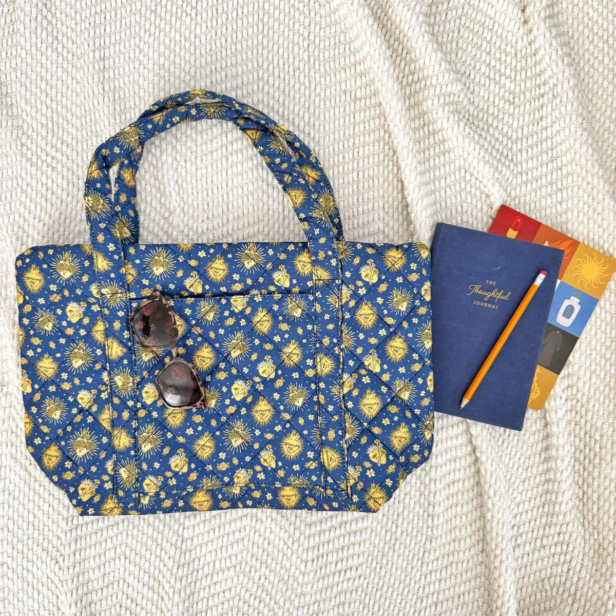 Holy Hearts Quilted Bag in blue with yellow pattern, spacious and versatile, featuring exterior pocket, sunglasses, and journal on white fabric.