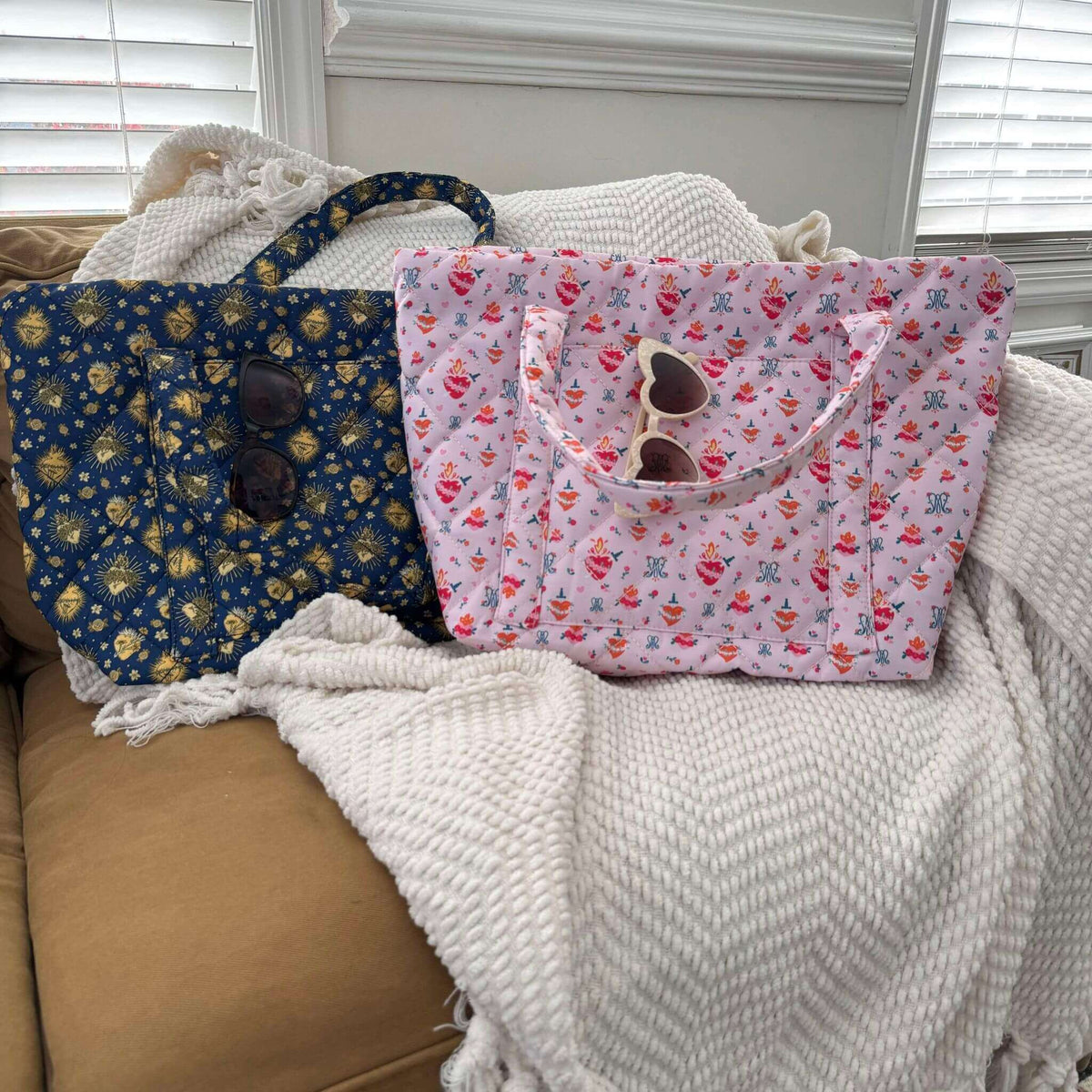 Holy Hearts Quilted Bags in pink and blue with patterns, lying on a cozy sofa with sunglasses in pockets. Spacious, stylish design.