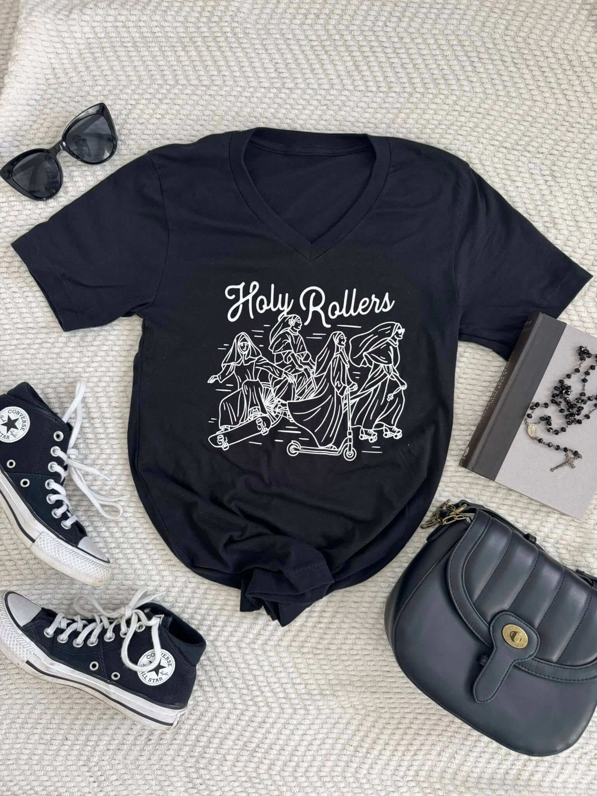 Holy Rollers Tee featuring nuns on scooters design, laid flat with black sneakers, sunglasses, bag, and book.