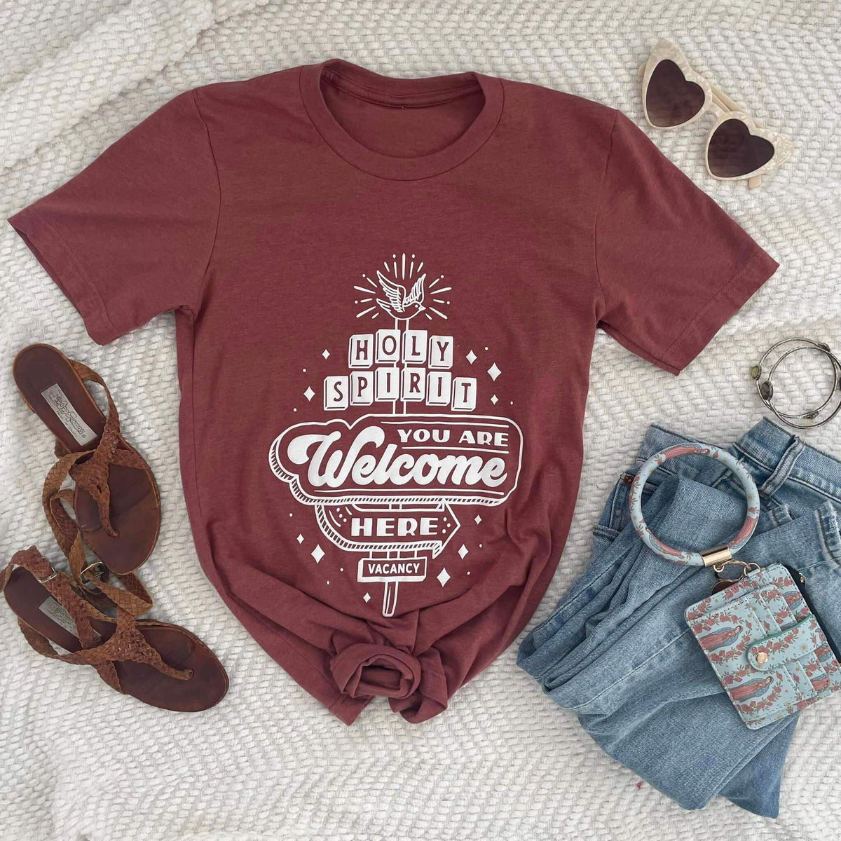 Holy Spirit You Are Welcome Here tee with vintage motel sign design, styled with jeans, sandals, watch, and heart-shaped sunglasses.