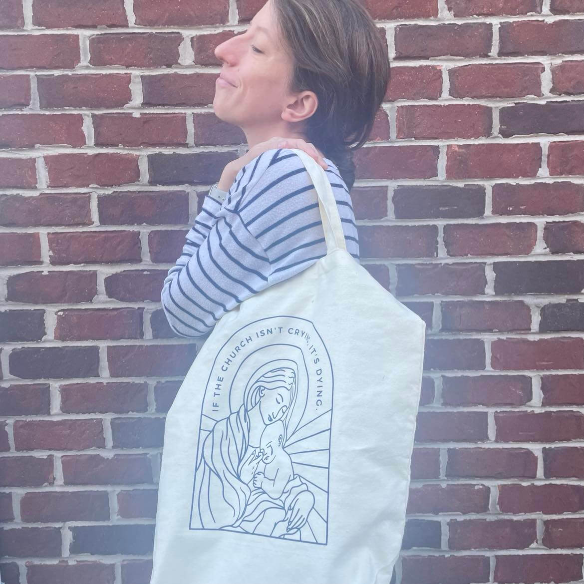 Woman holding a canvas tote bag with "If the Church Isn't Crying, It's Dying" message, featuring adjustable shoulder strap.