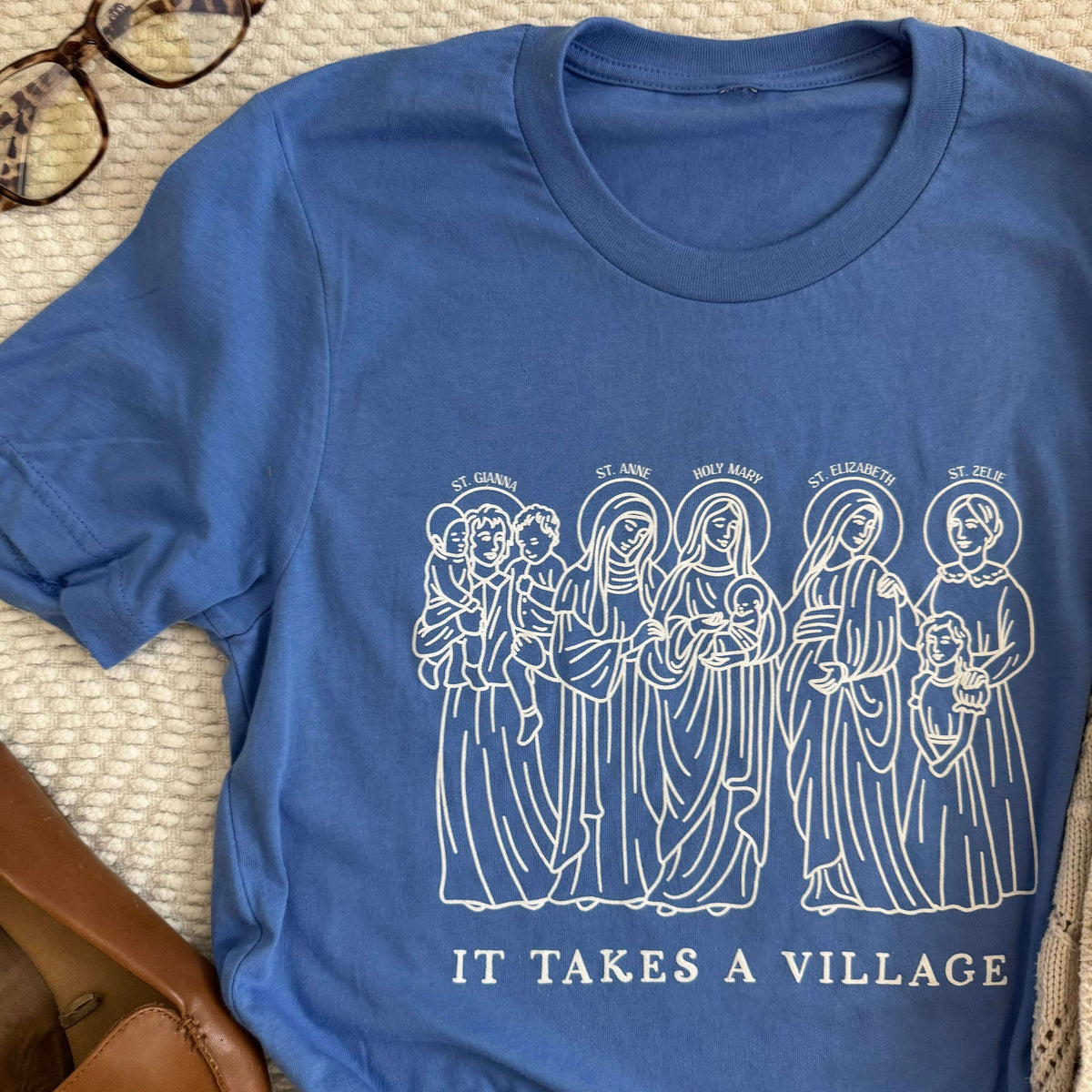Blue 'It Takes a Village' Tee with illustrations of St. Gianna, St. Anne, Mary, St. Elizabeth, and St. Zelie, spiritual motherhood theme.
