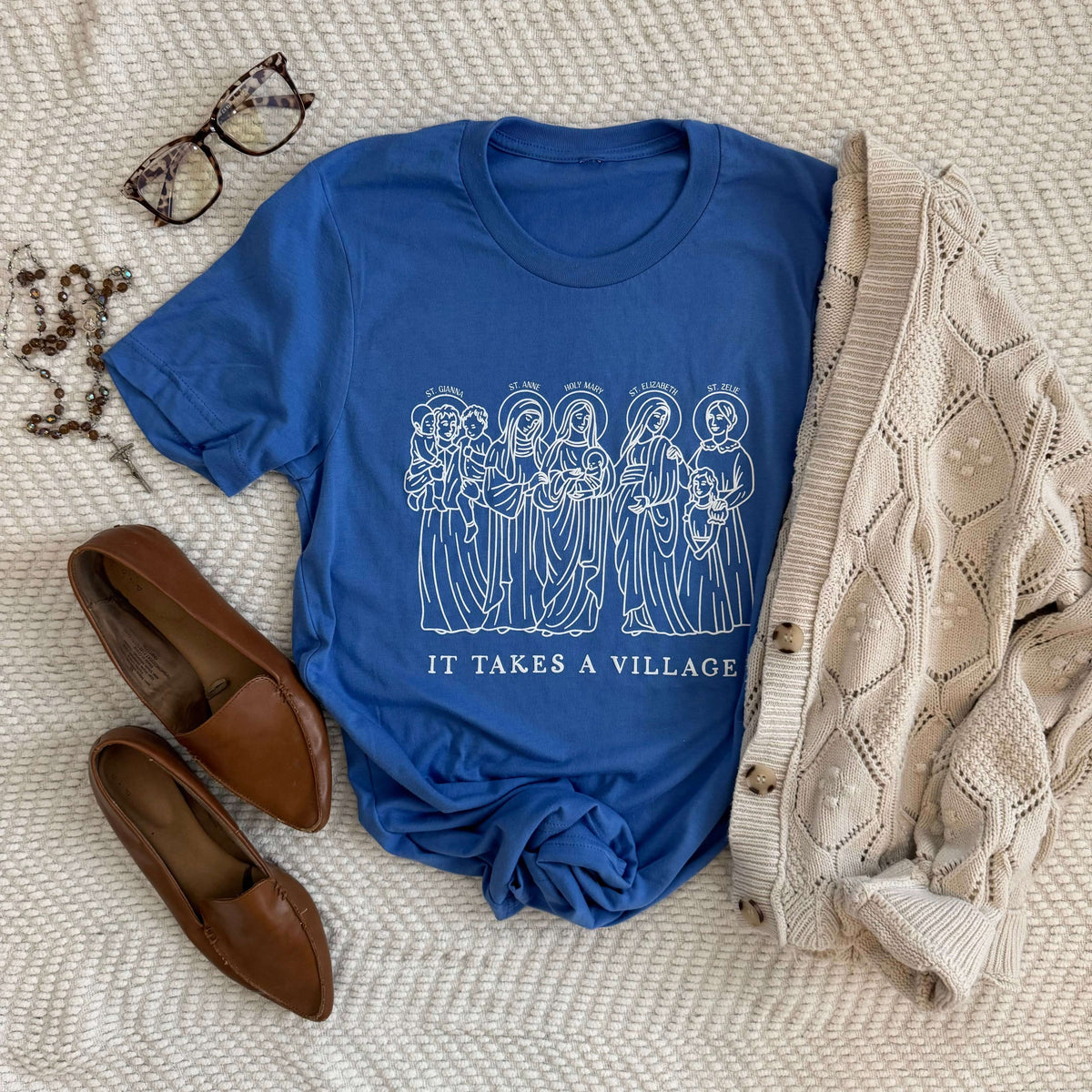Blue "It Takes a Village" tee featuring saintly mothers, styled with cardigan, shoes, glasses, and necklace on textured surface.