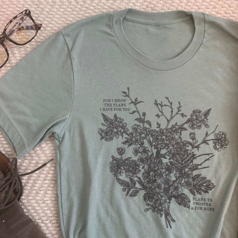 Plans to Prosper t-shirt featuring Jeremiah 29:11 with floral design, green background, laid beside glasses on textured surface.