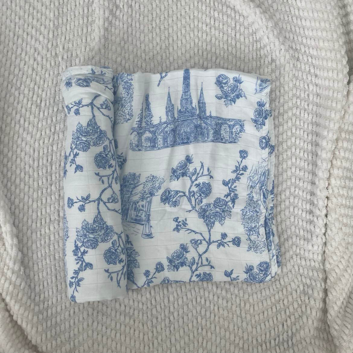 Lourdes Swaddle Blanket featuring blue floral and spiritual patterns, 100% organic bamboo cotton, 47x47 inches, on a textured surface.