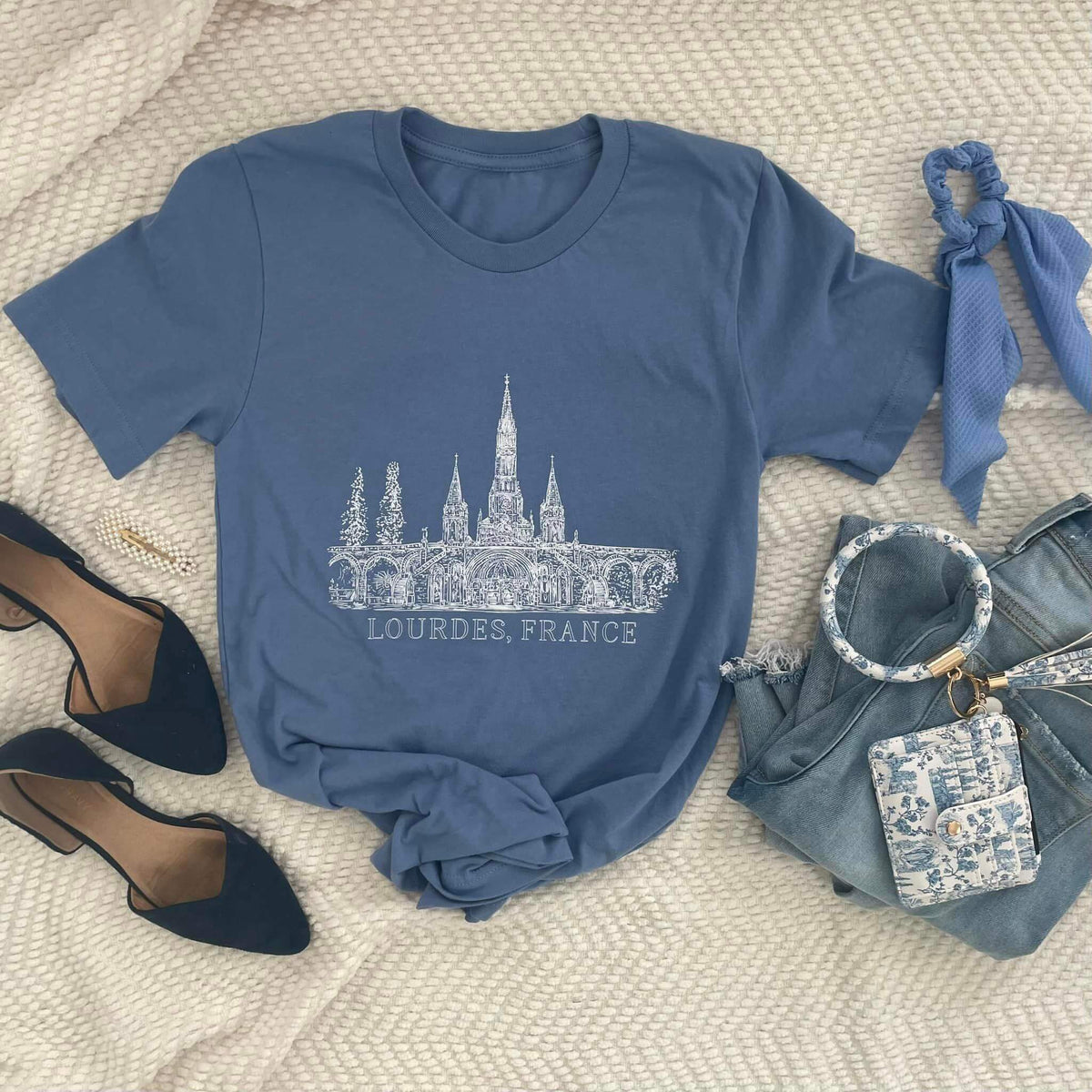 Blue Lourdes tee with Basilica Toile de Jouy illustration, featuring 'Lourdes, France' text, styled with accessories on a beige background.