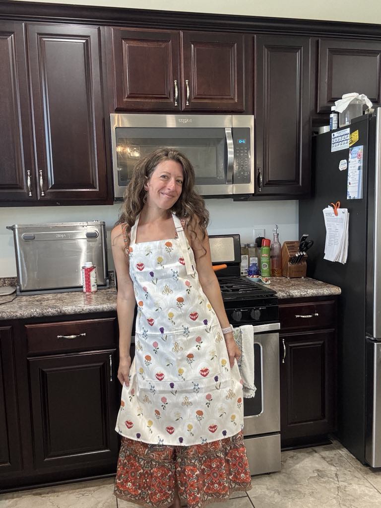 Woman wearing Mary's Sacred Garden Apron in a kitchen, featuring floral pattern and adjustable neck strap, promoting peace and functionality.