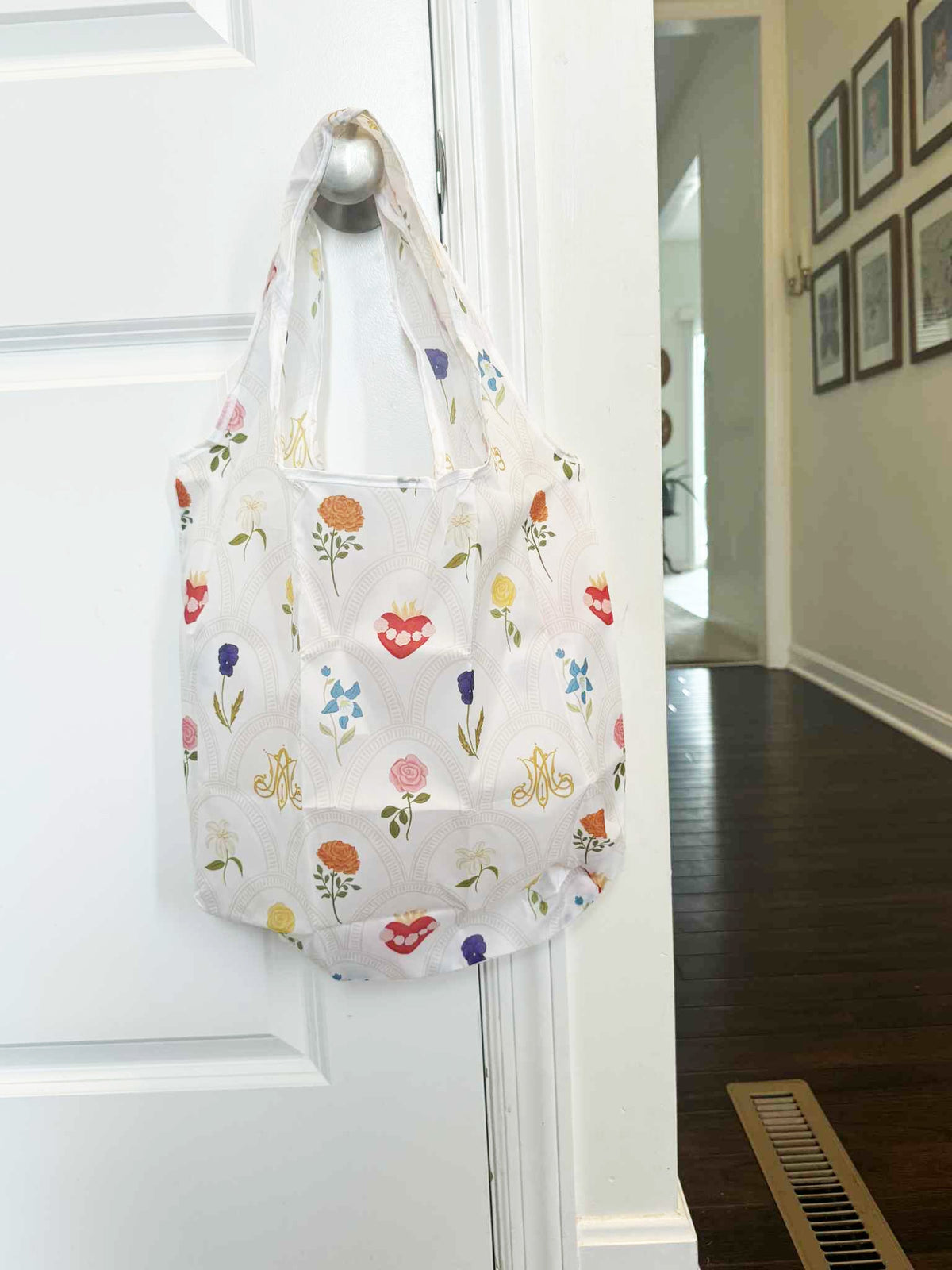 Catholic reusable grocery bag with floral design, part of Mary's Garden Collection, hanging on a door knob.