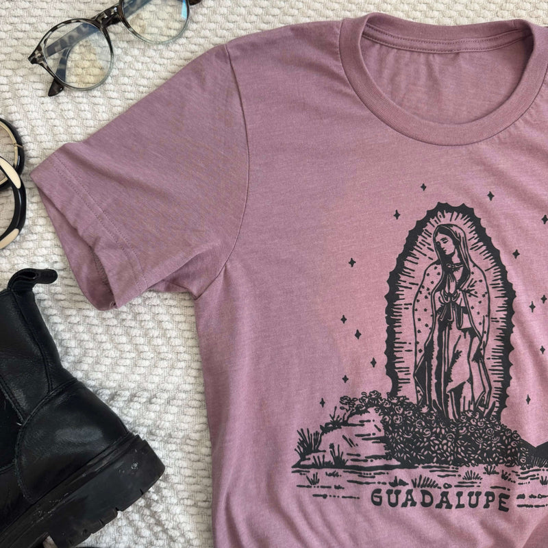 Pink Our Lady of Guadalupe t-shirt featuring a black print design, laid flat with black boots and glasses on a textured white surface.