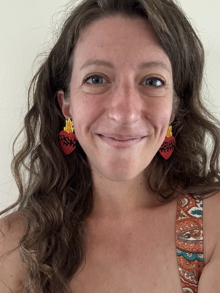 Woman wearing Sacred Heart beaded earrings, vibrant red and yellow design, showcasing joyful, bold style and meaningful devotion.