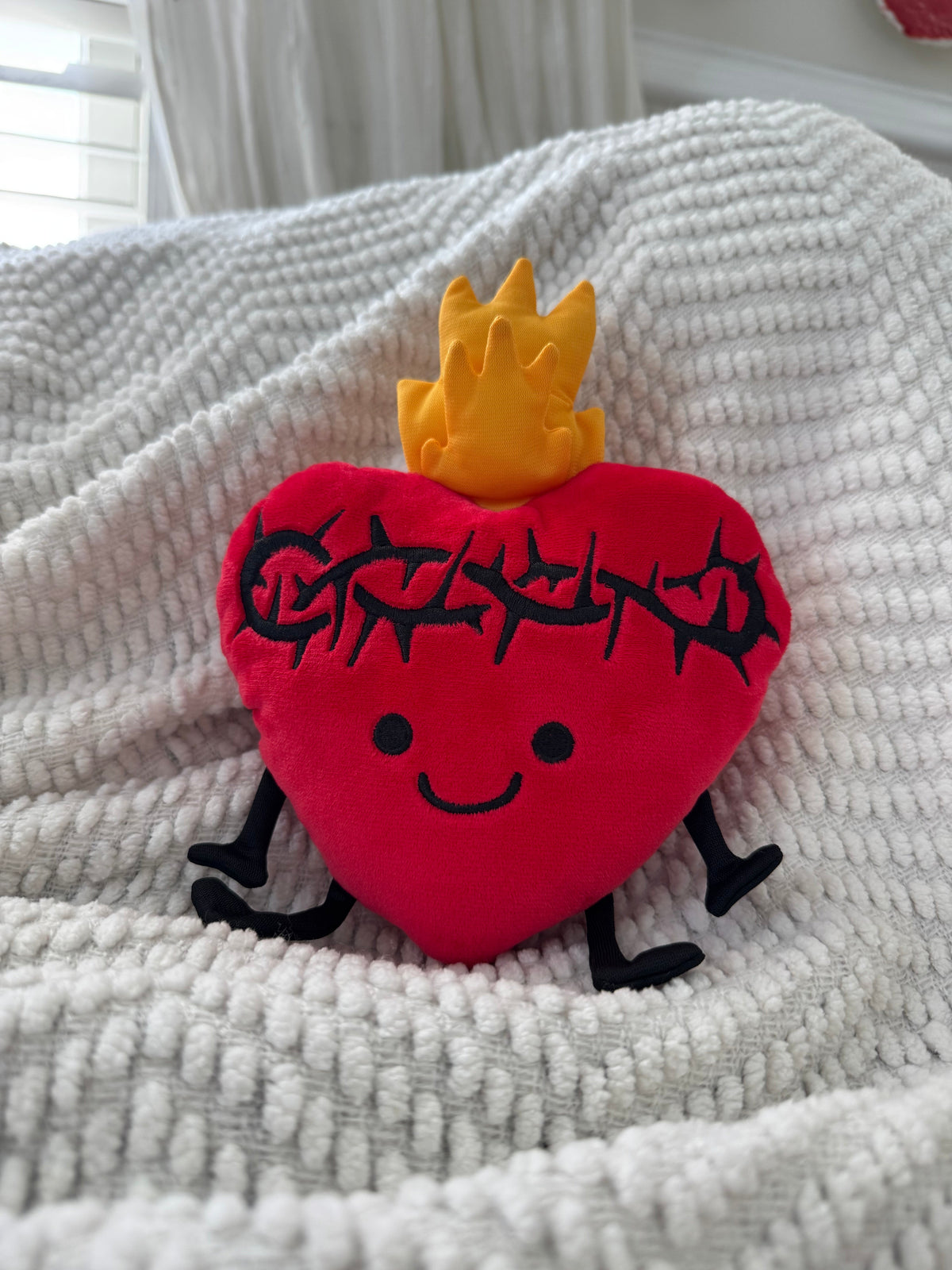Sacred Heart plush toy with joyful face and gentle flame, symbolizing love and comfort, on a cozy white blanket.