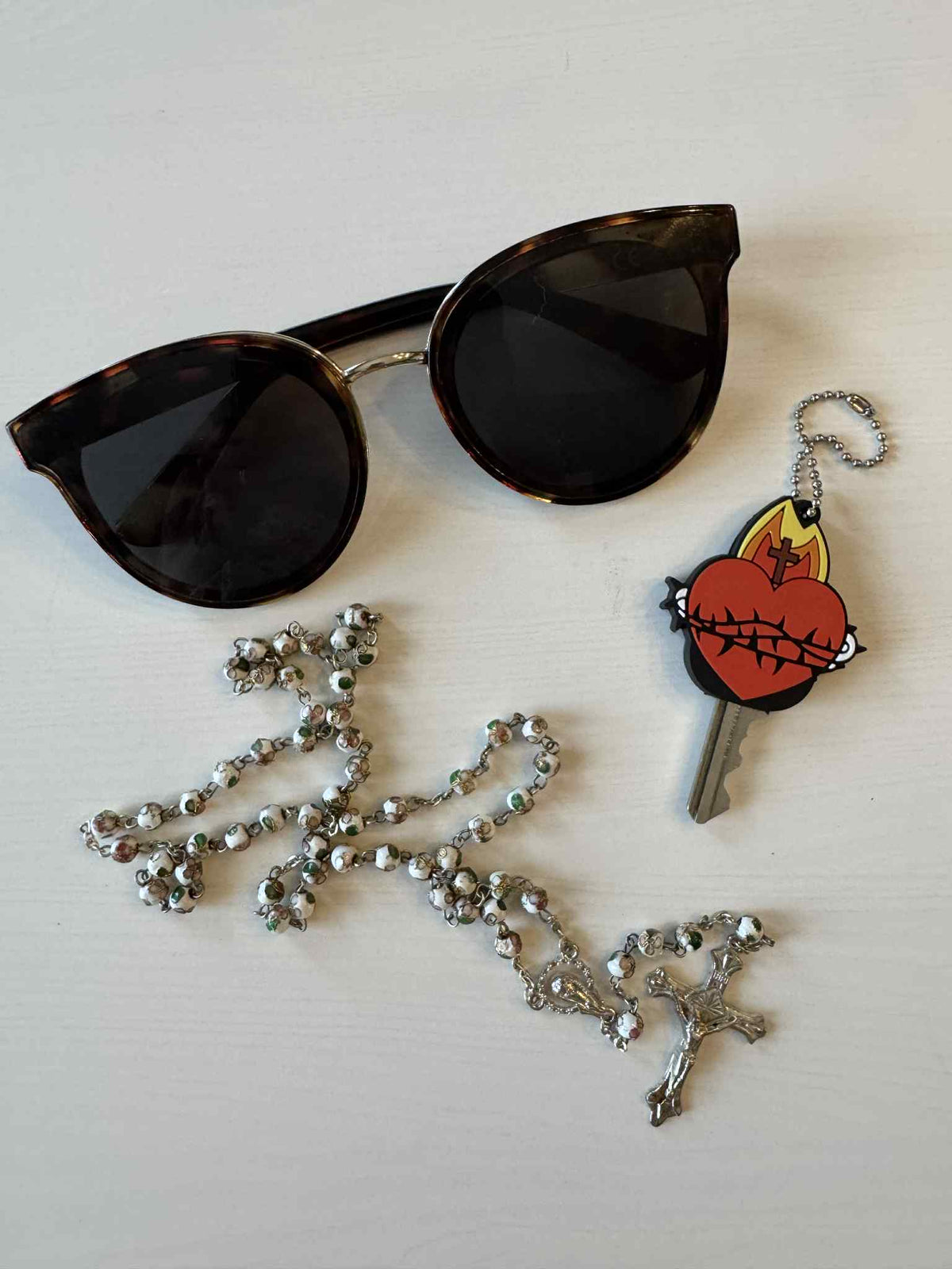 Sunglasses, rosary, and a key with a Sacred Heart silicone cap resting on a white surface, symbolizing devotion and protection.