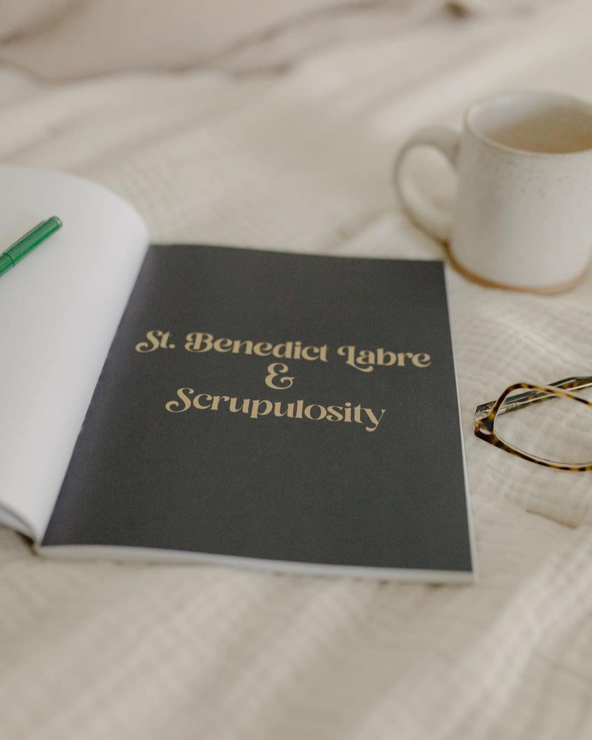 Open book on a bed displaying 'St. Benedict Labre & Scrupulosity', next to a coffee mug and reading glasses.