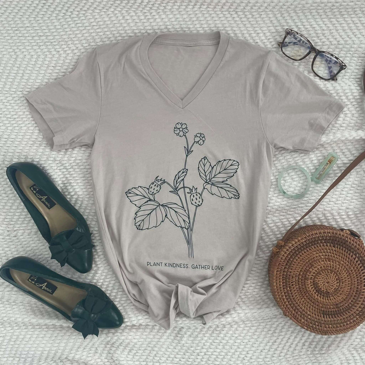 Saint Basil Tee with "Plant Kindness, Gather Love" design featuring strawberry plant, surrounded by accessories on white background.