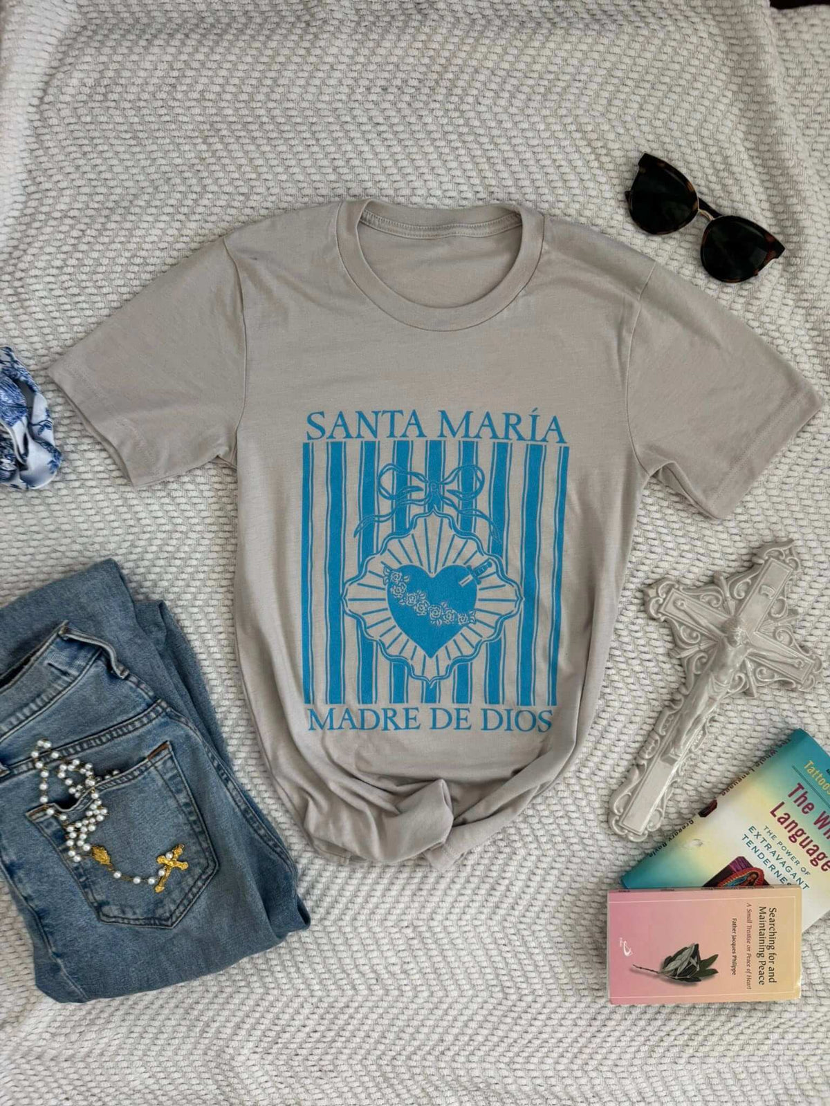 Santa María Madre de Dios T-shirt on a textured background with jeans, cross, sunglasses, and books. Features floral design.