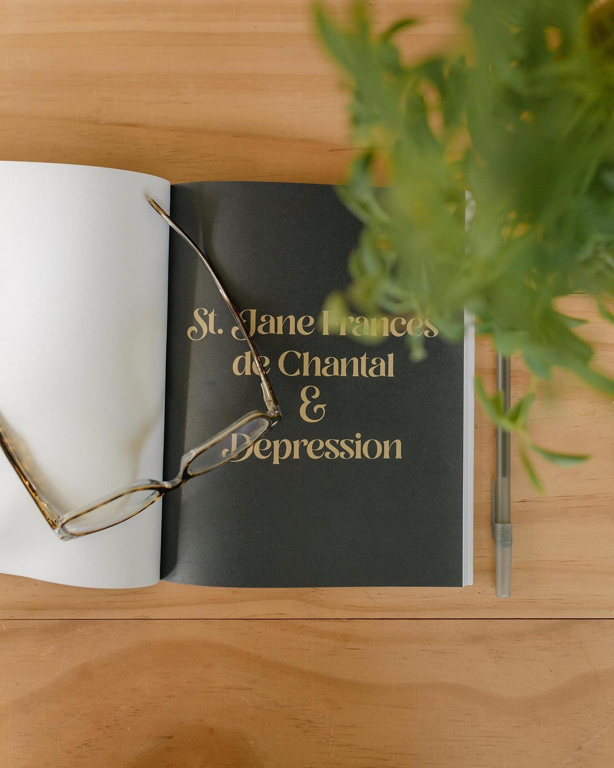 Open book titled 'St. Jane Frances de Chantal & Depression' with glasses and plant on a wooden table.