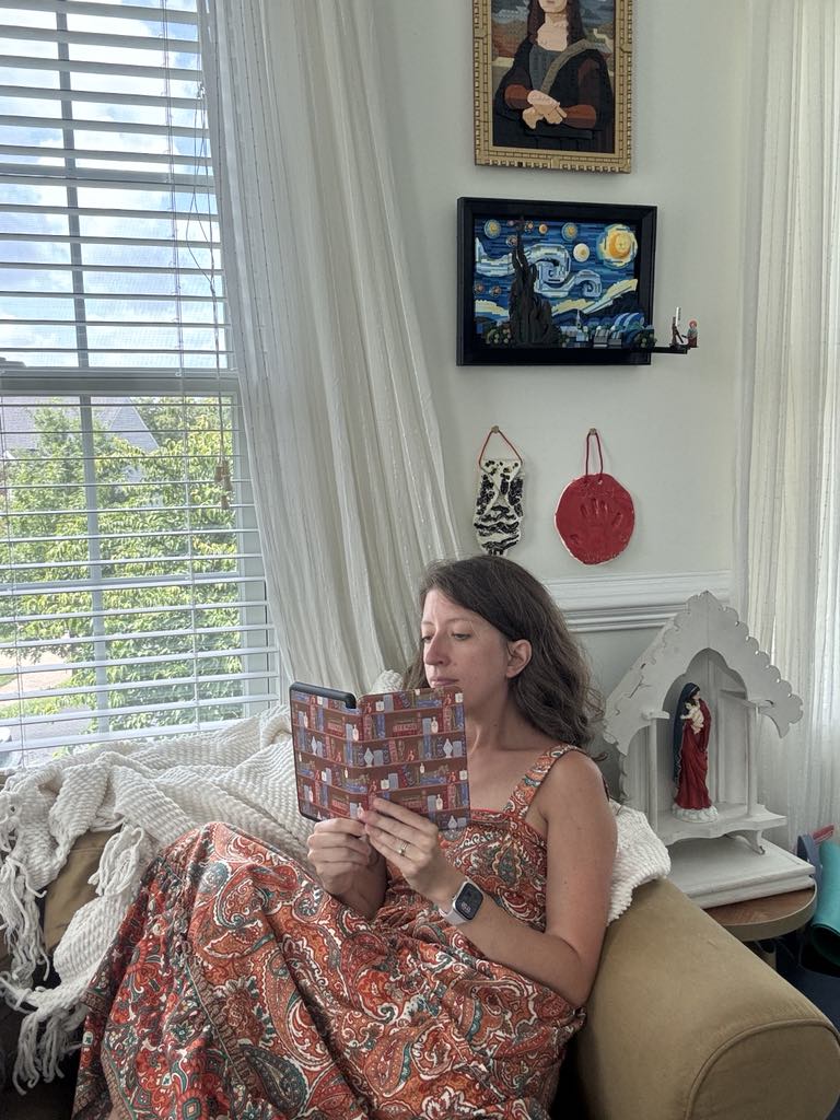 Woman reading Kindle with Catholic Library case, sitting in a cozy room with religious decor and art on walls.