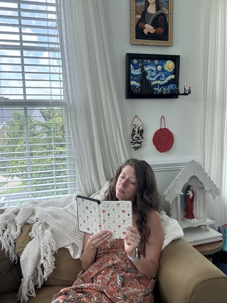 Woman reading on a couch with a Kindle in a floral 'Mary's Sacred Garden' cover, surrounded by religious decor.