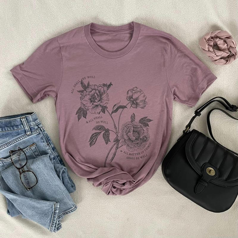Julian of Norwich 'All Shall Be Well' t-shirt with floral design, styled with jeans, glasses, and a black bag on beige background.