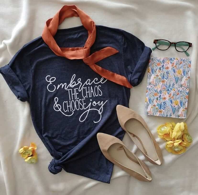 Heathered navy t-shirt with 'Embrace the Chaos & Choose Joy' slogan, styled with flowers, scarf, notebook, and glasses