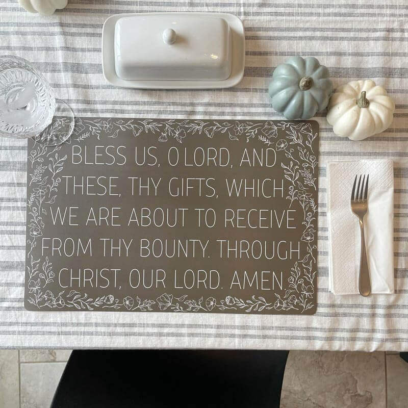 Grace prayer place mat on table with pumpkins, displaying the prayer 'Bless us, O Lord' in white ink on grey with floral borders.
