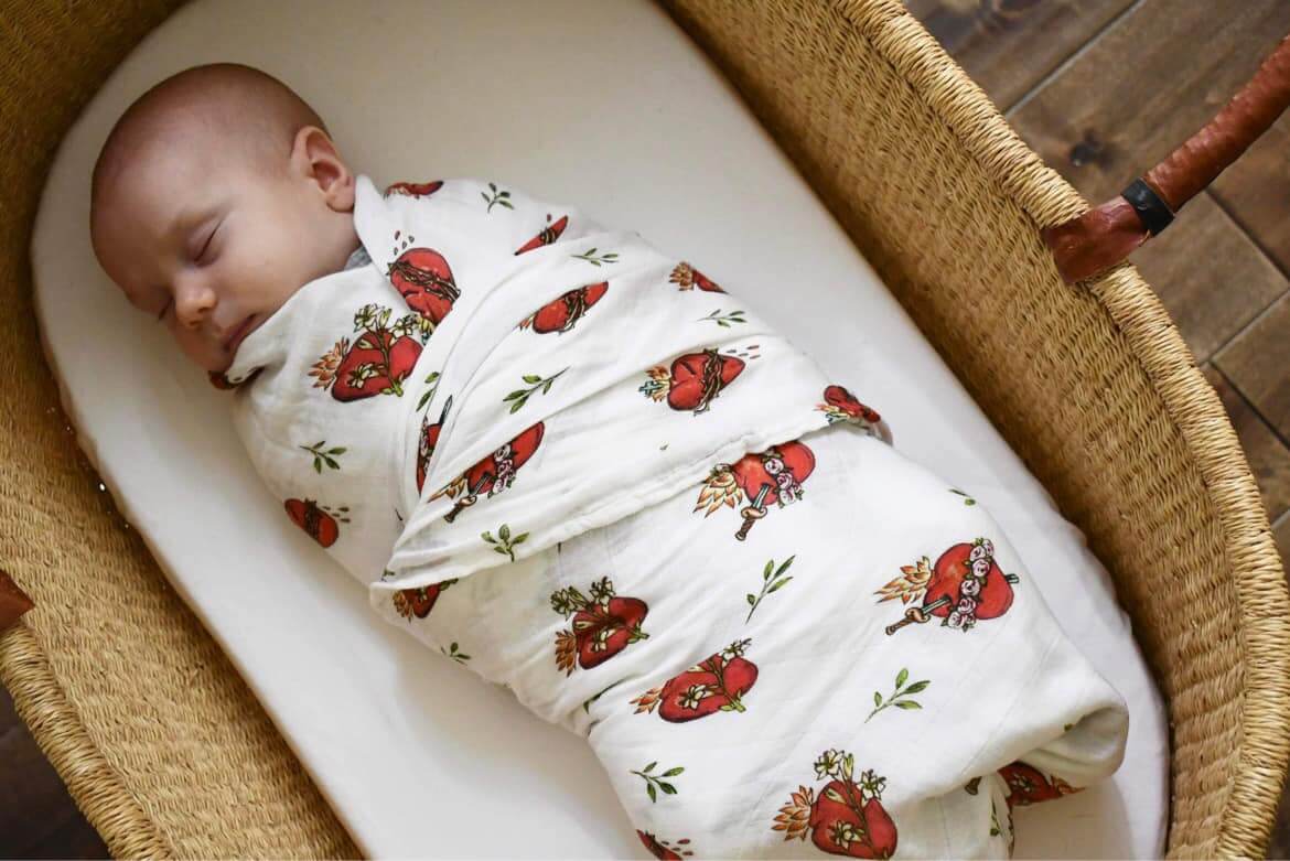 Baby sleeping in Holy Family Hearts Swaddle Blanket with sacred motifs in a wicker basket.