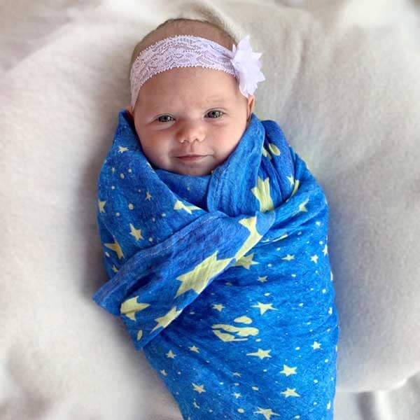 Baby wrapped in Mary's Mantle Swaddle, blue with yellow stars and wearing a floral headband, resting peacefully on a white blanket.