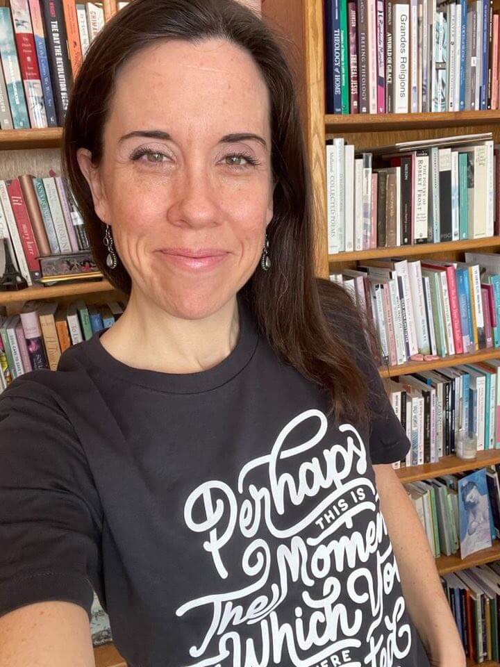 Woman wearing 'Perhaps This Is The Moment Tee' in front of a bookshelf, featuring quote from Esther 4:14 promoting self-reflection.