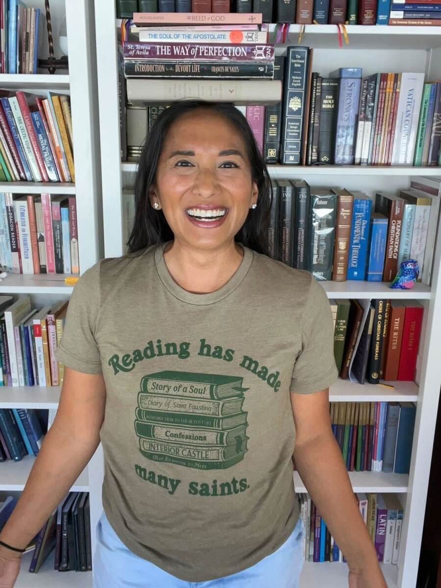 Woman wearing 'Reading Has Made Many Saints' St. Josemaria Escriva tee, standing in front of a bookshelf filled with spiritual books.