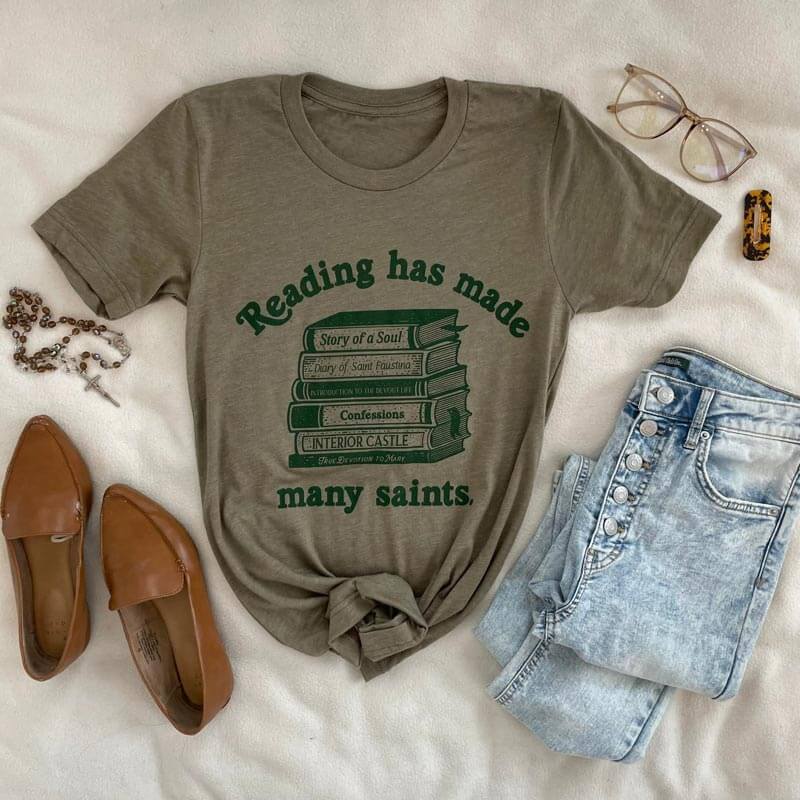 St. Josemaria Escriva tee with 'Reading has made many saints' quote, styled with jeans, brown shoes, and accessories.